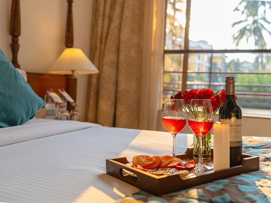 A close-up image of a bed featuring a tray that holds two half-filled wine glasses, a wine bottle, a candle, roses, and snacks.