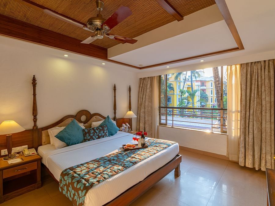An image of a room featuring a neatly made bed, a fan, two table lamps, side drawers, and a large window.