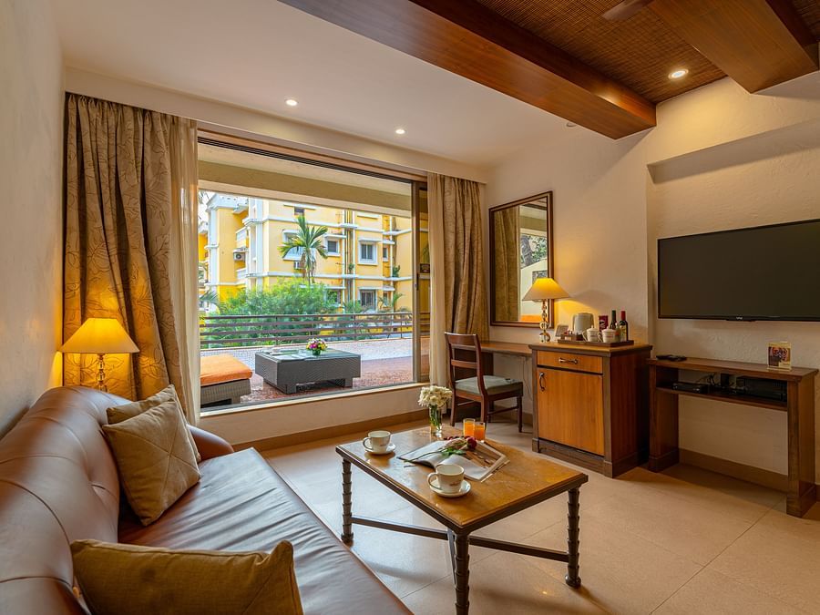 An image of a living room with a brown sofa, small coffee table, TV, storage cabinet, tea and coffee maker, and a balcony with a seating area.
