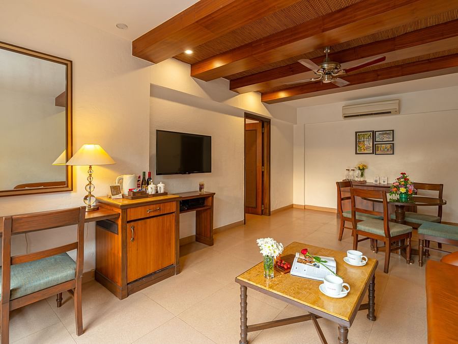 An image of a living room featuring a dining table and chairs, a coffee table, a mirror, a TV, a tea and coffee maker, wall paintings, and an AC unit.