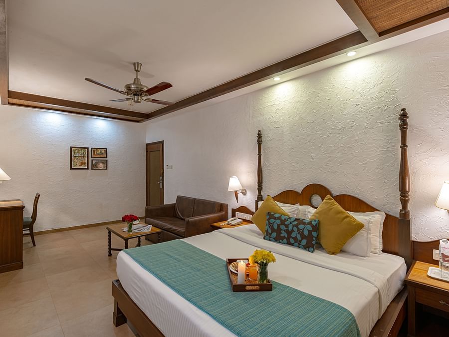 An image of a room with a sofa, table lamps, a vanity, wall paintings, and a neatly made bed featuring a tray with a candle, juice, flowers, and snacks.