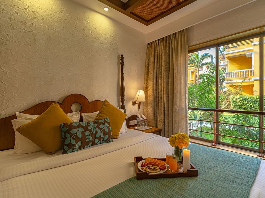 An image of a room showcasing a beautifully decorated bed adorned with a tray of candles, flowers, and fruits, complemented by a large window.