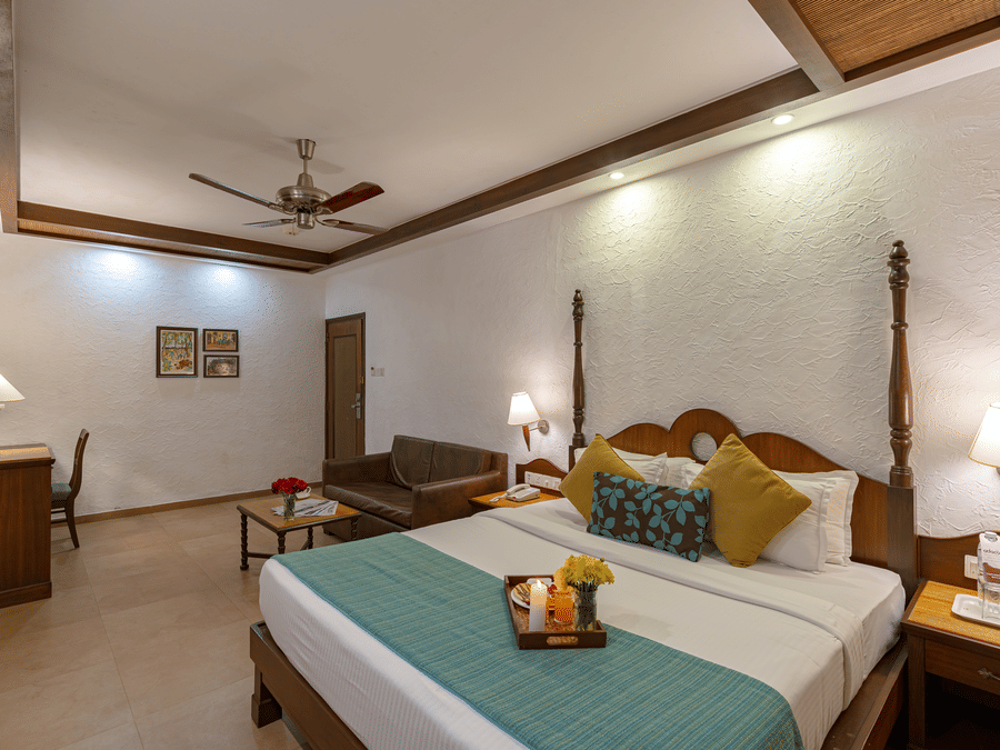 An image of a room with a sofa, table lamps, a vanity, wall paintings, and a neatly made bed featuring a tray with a candle, juice, flowers, and snacks.