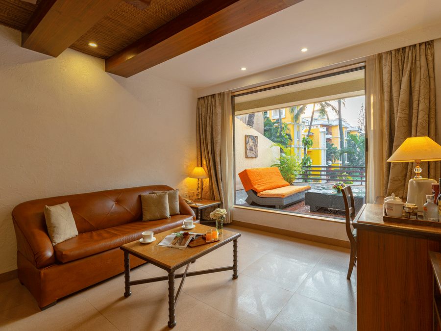 An image of a living room with a brown sofa, coffee table, table lamps, a tea and coffee maker, and an attached balcony with seating.