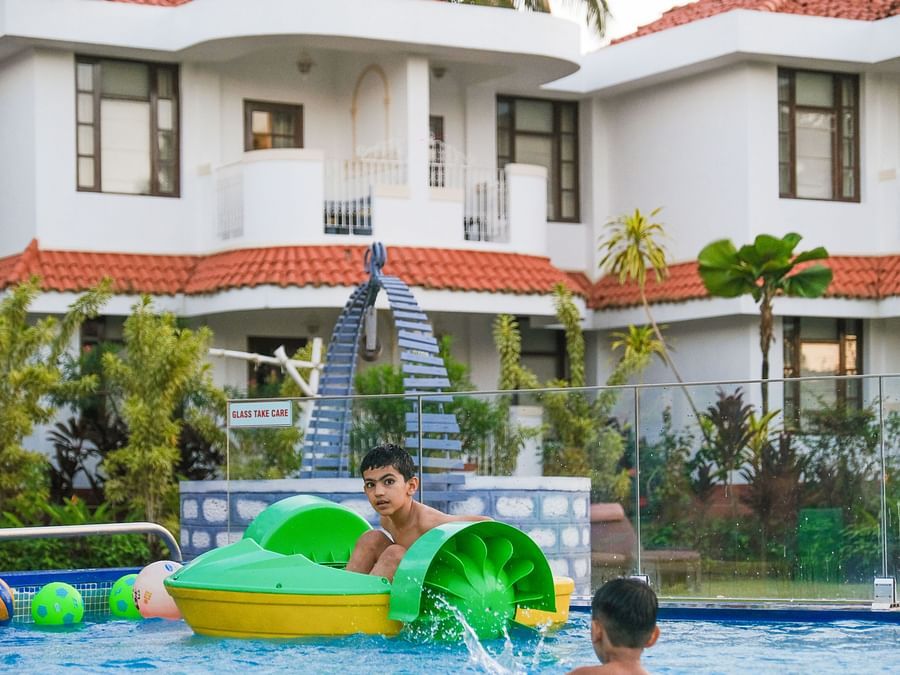 The two child enjoying in the swimming pool with the background of the big villa and tress at Heritage Village Resorts & Spa, Goa.