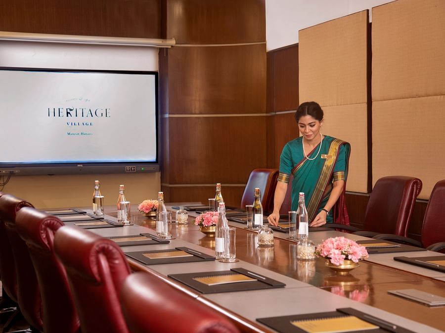 A modern boardroom setup featuring leather chairs, a wooden conference table with notepads, water bottles, floral decor, and a large presentation screen