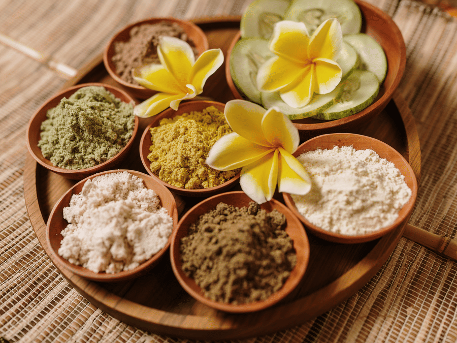 Assorted natural body scrubs and spa ingredients arranged in wooden bowls with tropical flowers.