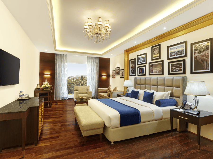 The executive suite bedroom with a king size bed, seating area, television, wooden flooring, framed wall decor and elegant lighting at La Marvella, Bengaluru.