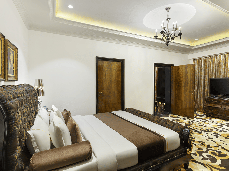 The spacious presidential suite bedroom with a king size bed, upholstered headboard, bedside tables, chandelier lighting, television unit and patterned flooring at La Marvella, Bengaluru.