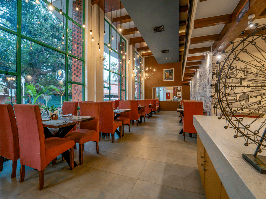 Rosetum Anjuna's ground floor restaurant with terracotta high-back chairs, wooden tables, Edison bulb pendant lights, floor-to-ceiling green-framed windows, and a mural wall.