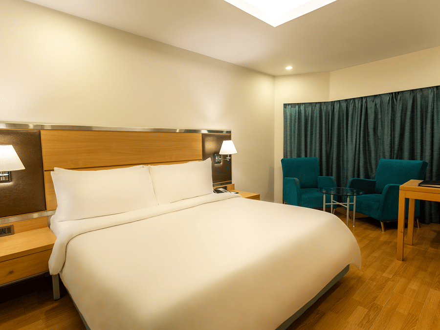 Superior Panoramic Rooms at The Residency Chennai featuring a plush bed with a wooden headboard and lamps on both sides above the nightstands, a sitting space of two couches and a coffee table by the window