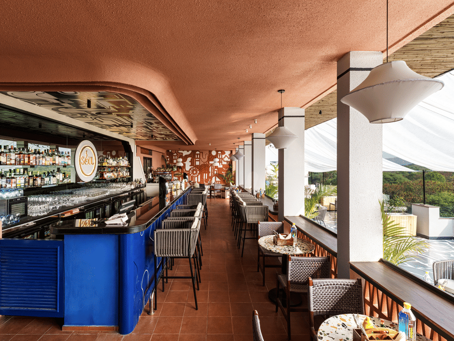 A long rooftop bar counter at The Sierra - By The Lake features stools, small dining tables, and hanging lamps under a shaded structure with open sides.