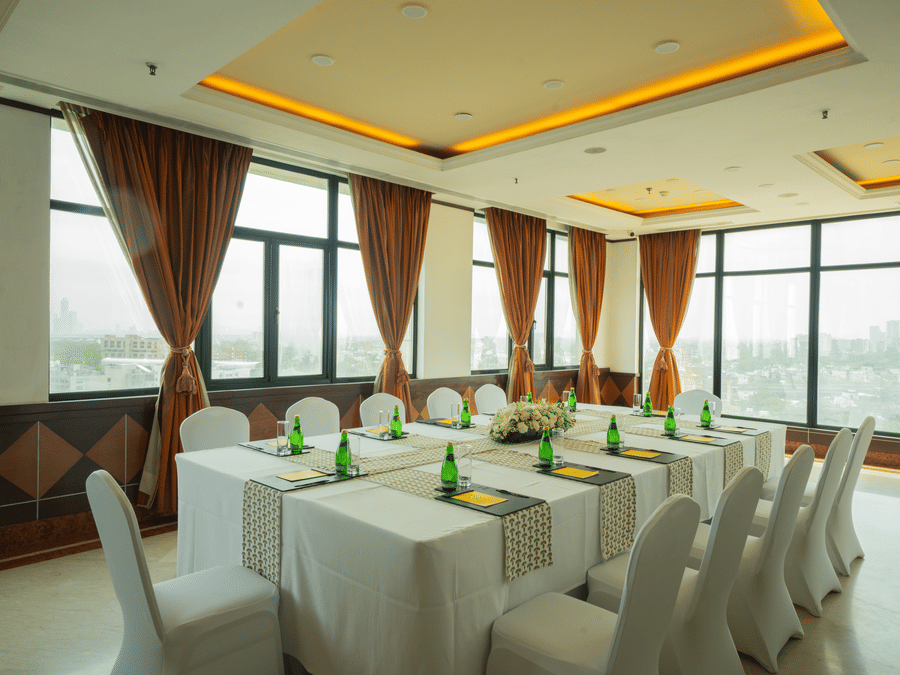 Sampan Tea Room at The Suryaa set with rectangular tables, white covers, green bottles, notepads, pens, warm recessed lighting, large windows with city view and brown curtains.