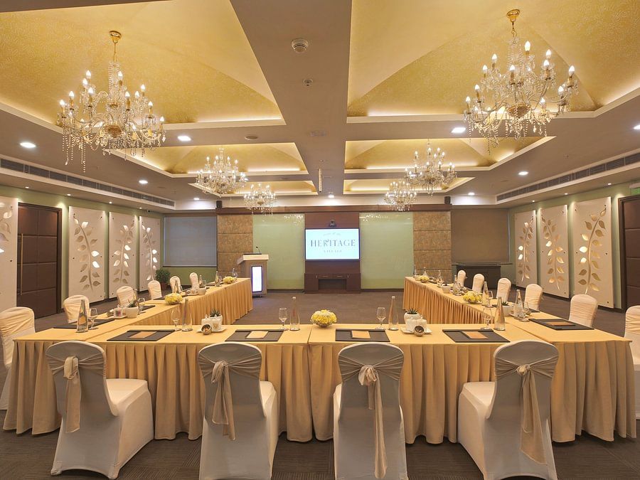 Well-lit conference room with a U-shaped table, elegant chandeliers, and a screen displaying HERITAGE - Heritage Village Resorts & Spa.