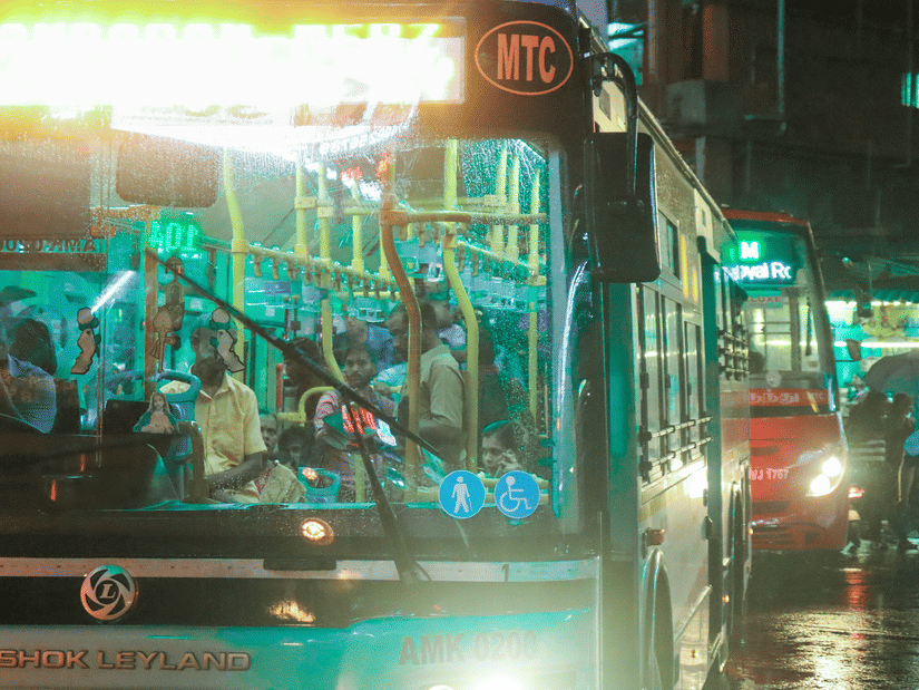 Bus at night with bright headlights, passengers visible inside, wet streets reflecting lights, and urban buildings glowing in background