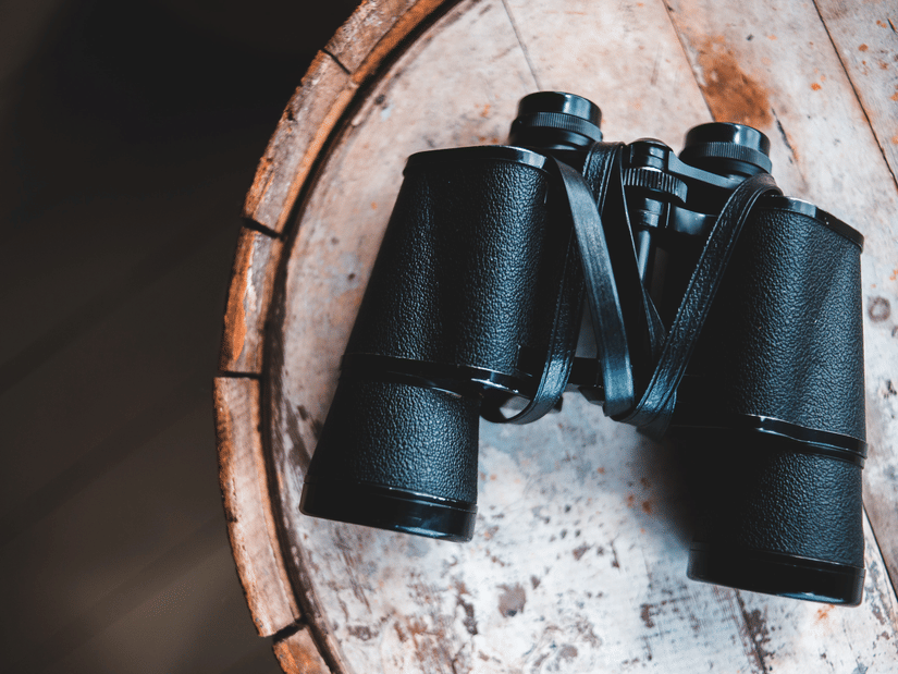 Black binoculars placed on a round wooden surface, with textured tabletop and dark background visible around the object.