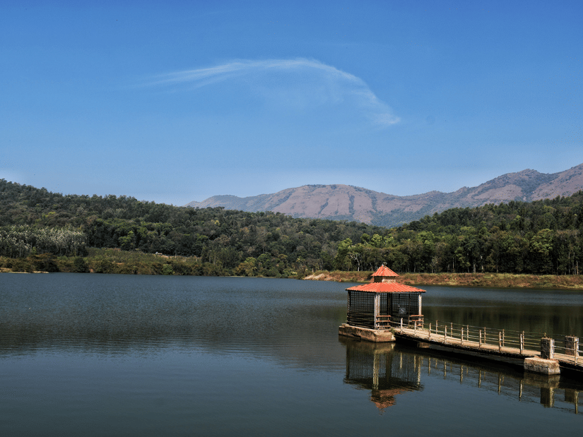 Peaceful lake with a wooden jetty and gazebo, surrounded by lush greenery and hills beneath a clear blue sky.