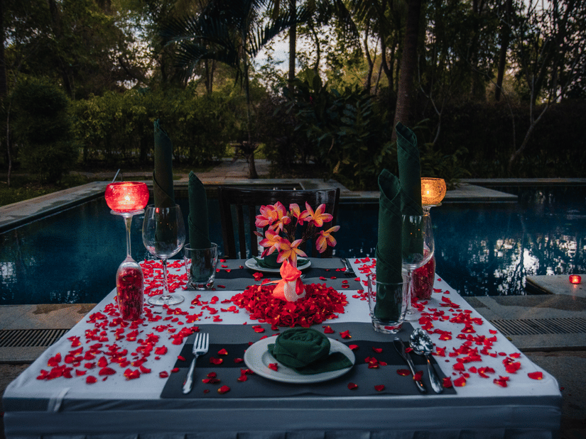 A dinner table set by the pool at The Serai Chikmagalur with rose petals and candles around the cutlery
