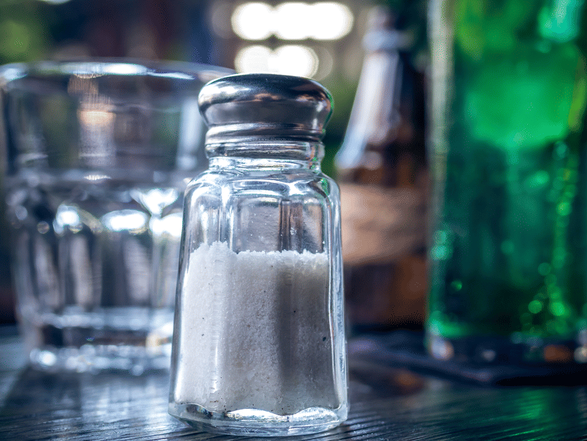 A clear glass salt shaker with a silver top sits on a dark table next to a glass and a bottle.