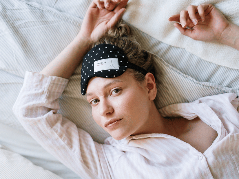 A woman lying in bed with her hands over her head and a sleeping mask on her forehead