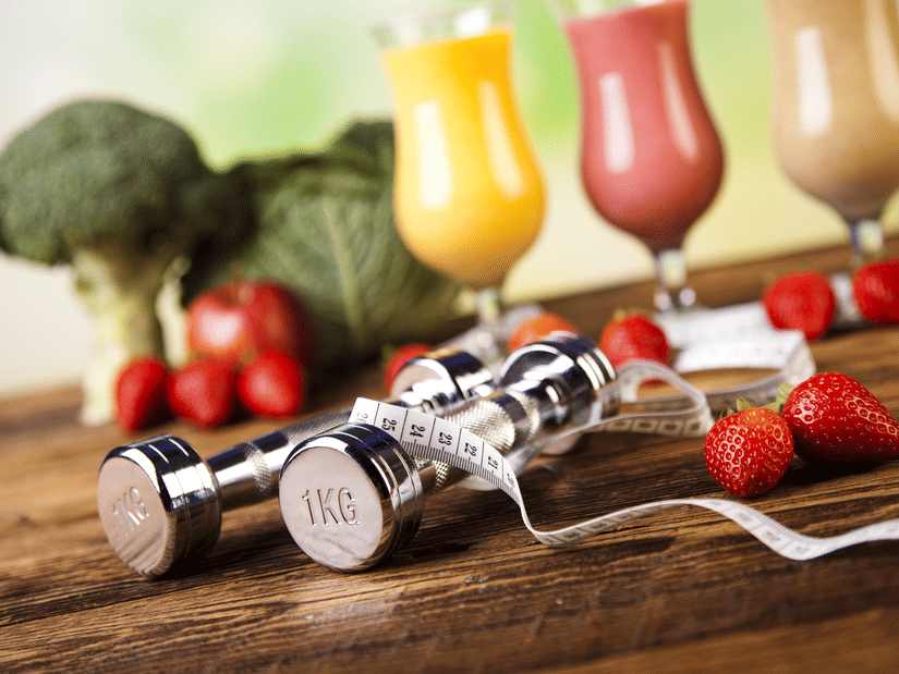 A set of 1 kilo dumbbells on a wooden surface with a set of vegetables and glasses of juices in the background