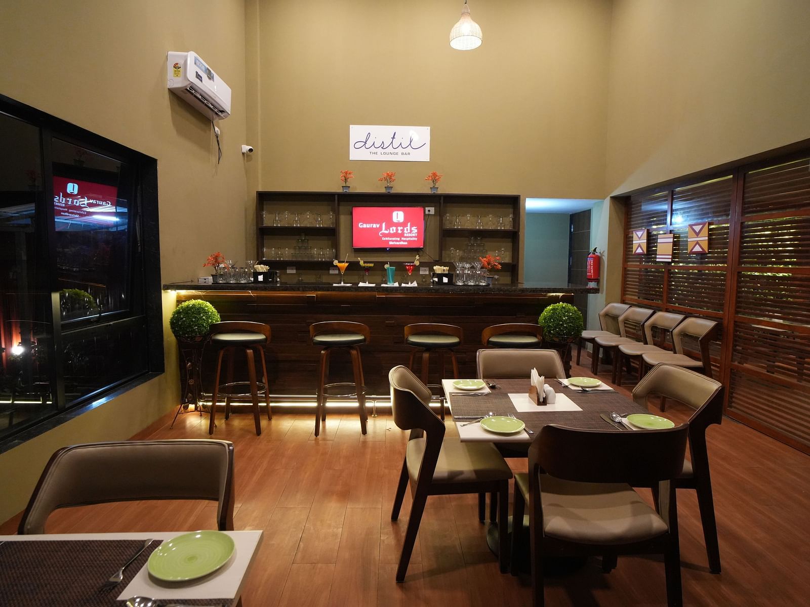 A cosy lounge bar with wooden flooring, high stools, cocktail displays, dining tables set with green plates, and large windows showing city lights at night - Gaurav Lords Resort Shrivardhan