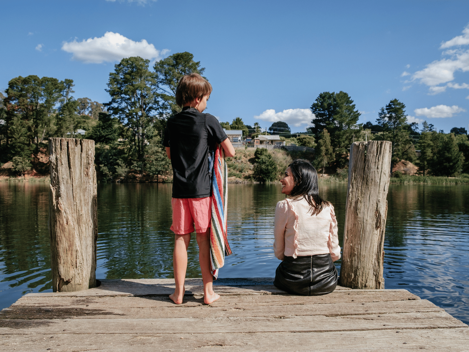 A young boy and girl sitting on a wooden pier overlooking a calm lake surrounded by lush greenery under a bright blue sky.