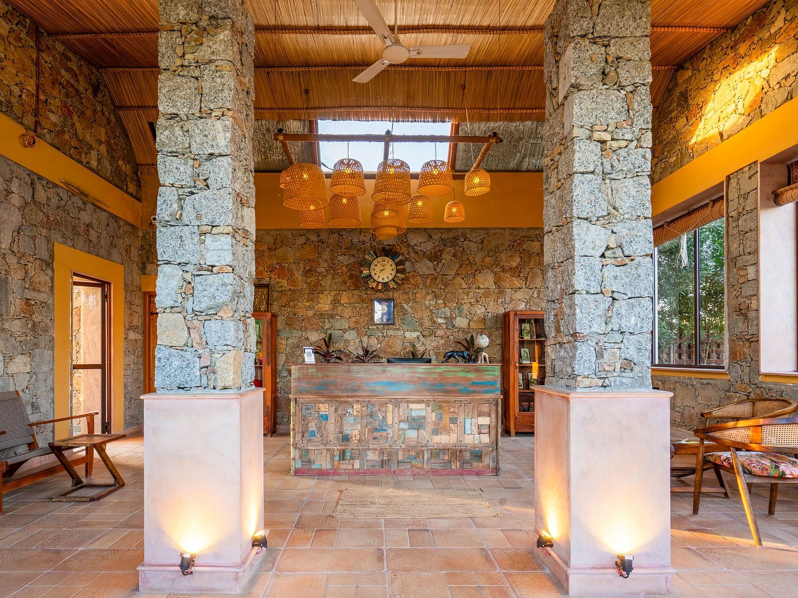 An image of a reception in bohemian theme and rock pillars and walls featuring  wooden seating area and a reception desk, and above it there are bamboo lamps - Kurja Jawai