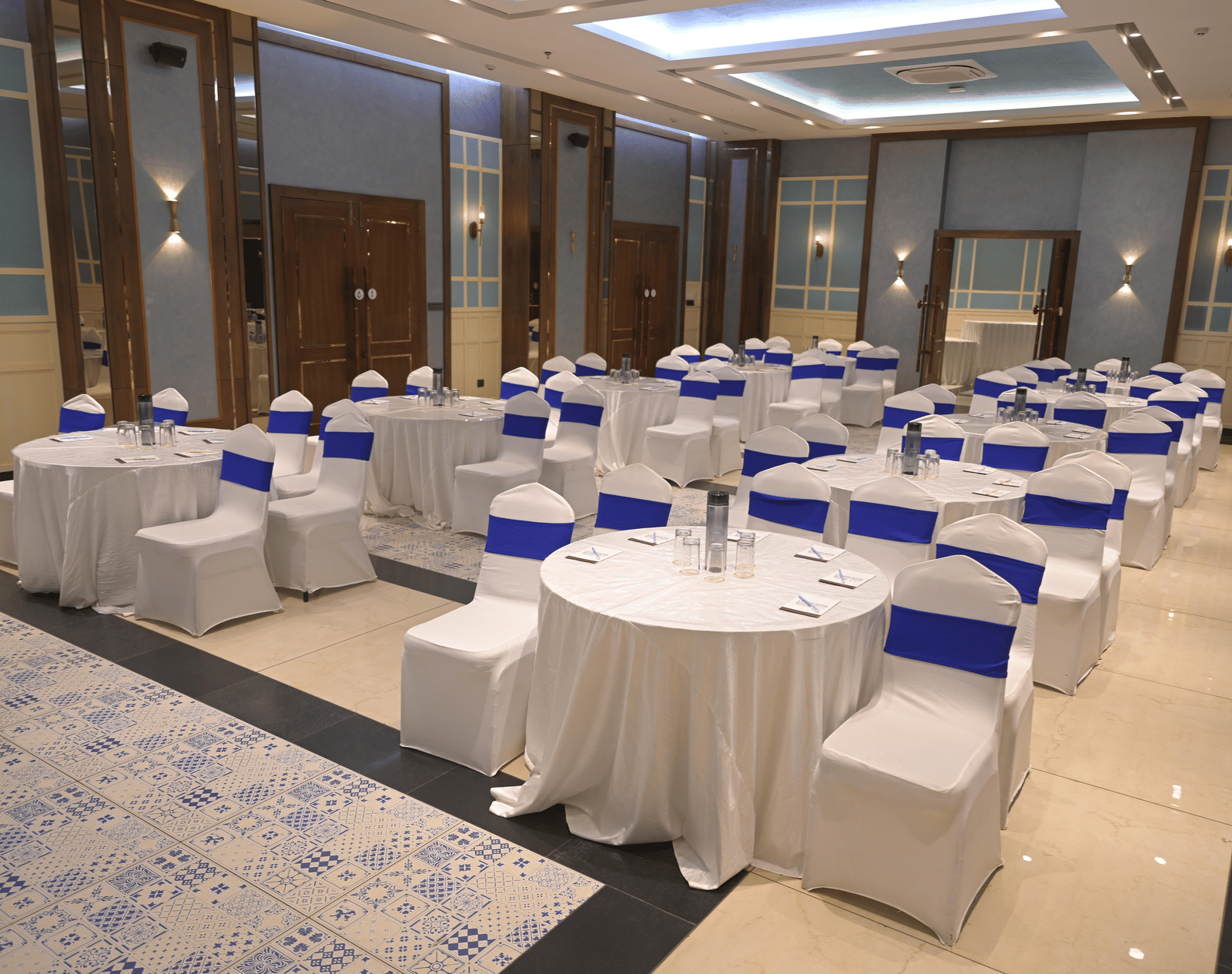 Round banquet tables arranged with white covers and blue chair sashes in a spacious event hall at Hotel Sonar Bangla Mayapur.