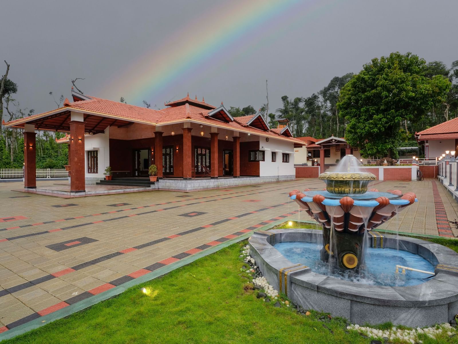 The traditional facade of Bhuthanakadu Retreat under a vibrant rainbow, featuring a fountain in a garden and a colorful patterned walkway leading to the main buildings.