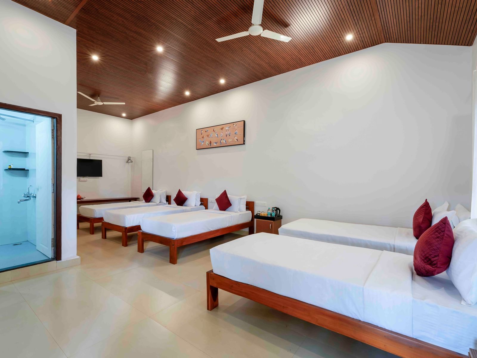 A spacious hall of residences at Bhuthanakadu Retreat, showcasing four single beds with white bedding and red pillows, a modern bathroom, and a warm wooden-paneled ceiling.