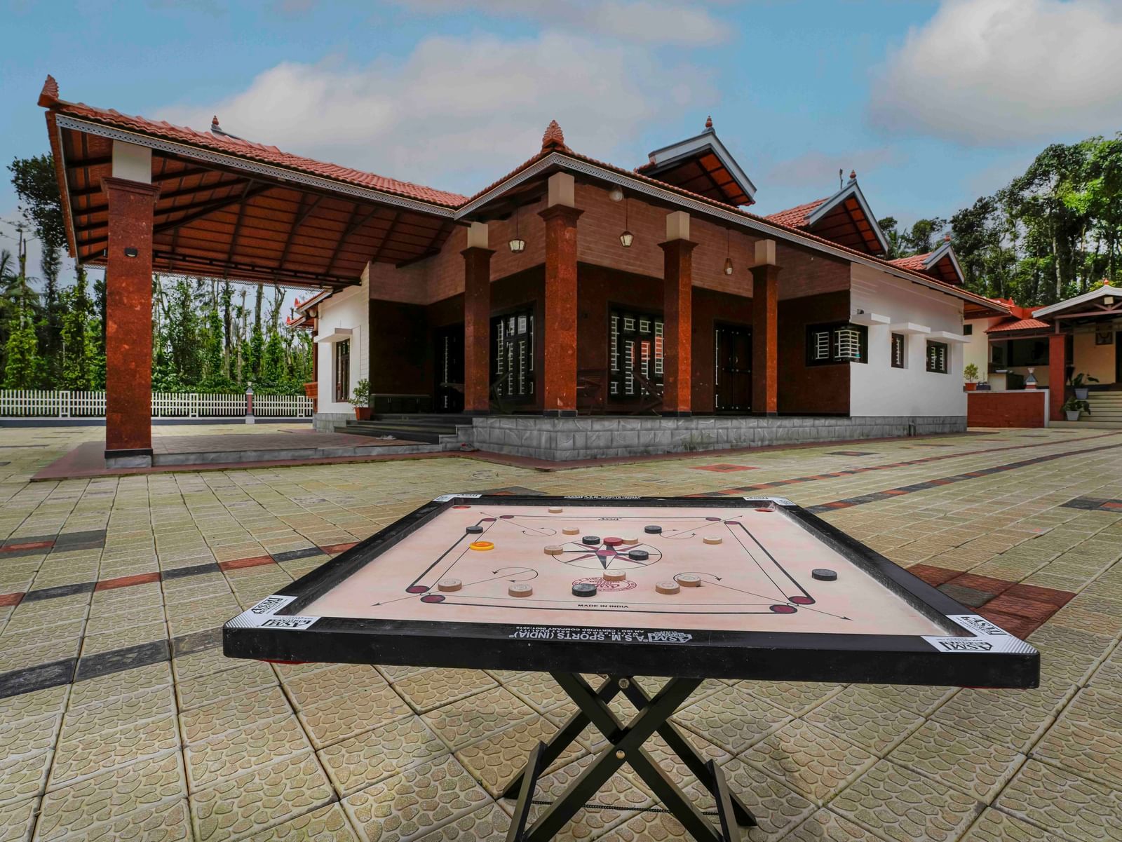 A carrom board is set up outdoors on a paved courtyard, with the traditional architecture of Bhuthanakadu Retreat visible in the background.