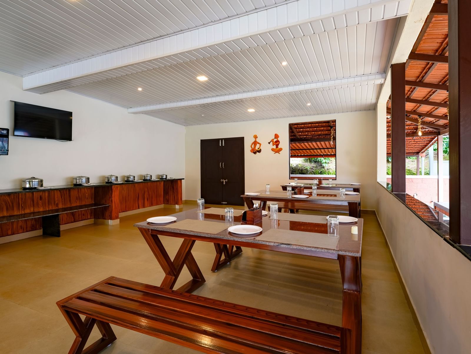 A daytime interior view of the Mane Cafe at Bhuthanakadu Retreat, highlighting a buffet area with covered dishes, a TV on the wall, and long wooden dining tables with benches.