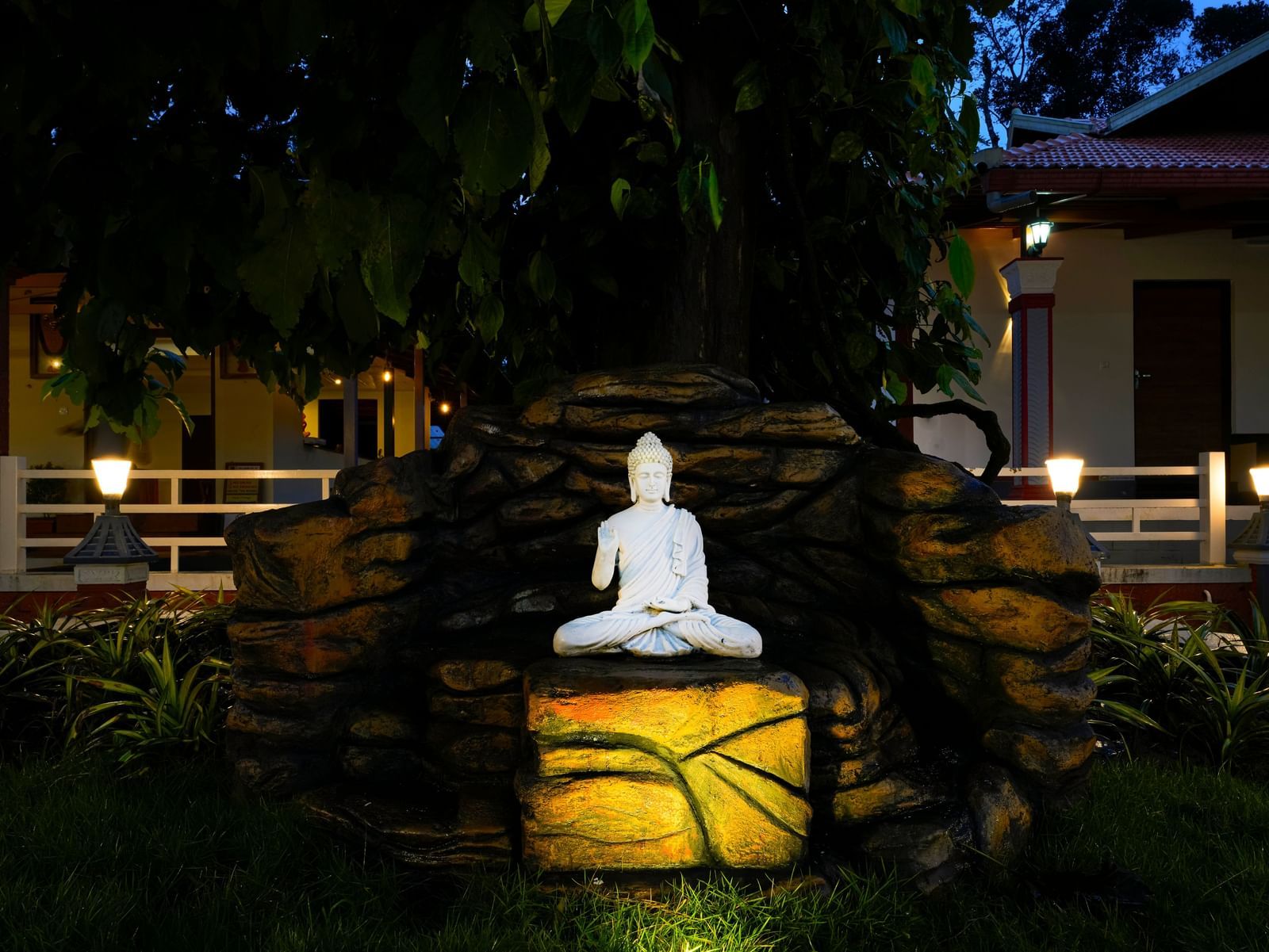 A serene Buddha statue sits on a rock illuminated by a soft, yellow light in a lush garden at Bhuthanakadu Retreat, with buildings visible in the background.