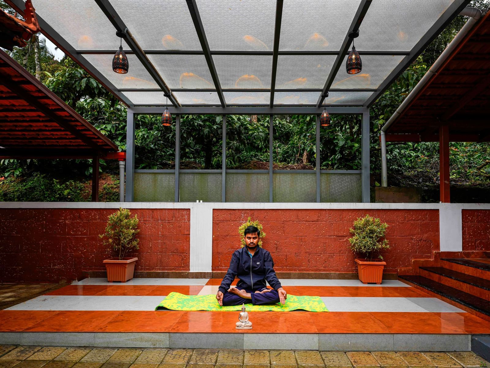 An individual is meditating in a serene, partially covered outdoor yoga space at Bhuthanakadu Retreat, surrounded by lush greenery and red-tiled walls.