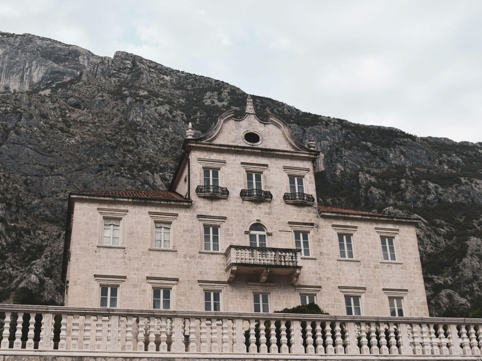 An old, majestic, multi-story building with a balcony and an arched entryway, set against a backdrop of a large mountain.