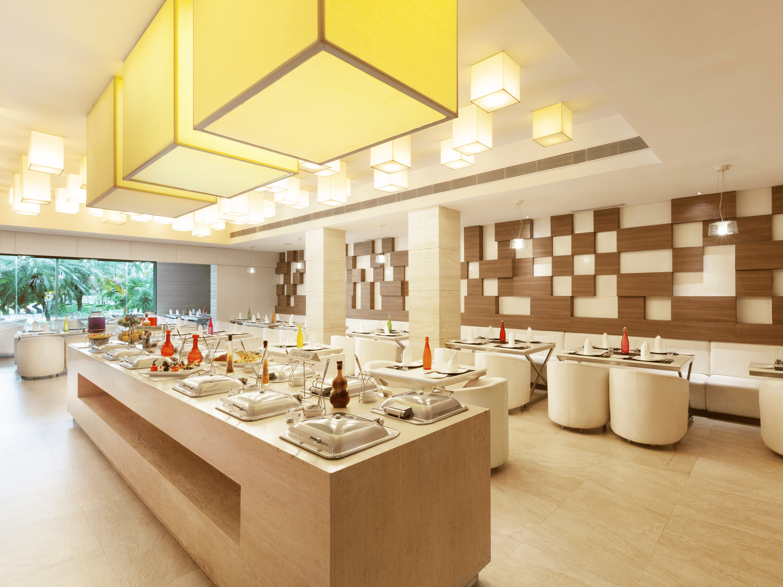 A diagonal view of a restaurant with an island in the middle featuring buffet items, surrounded by tables and chairs on the sides | Cenneys Gateway