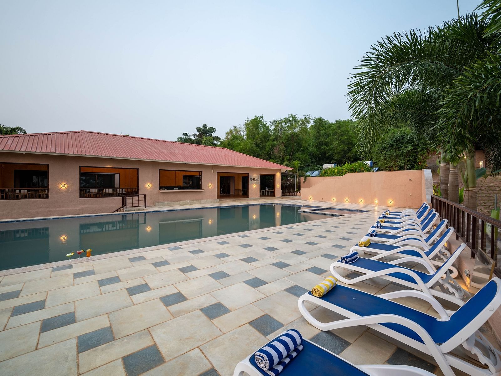 A poolside scene with beach chairs, a swimming pool, and a building framed by lush greenery on one side - Crescent Resort, Goa.