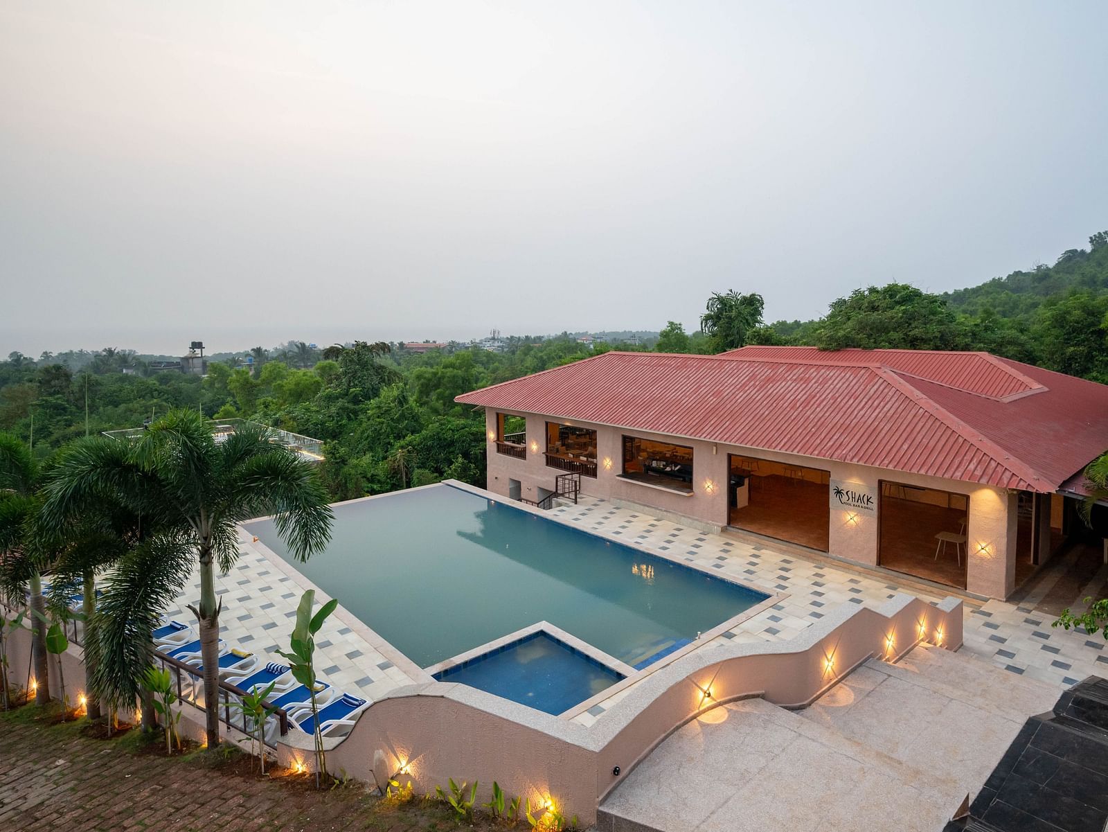 An eagle-eye view of a scene featuring beach chairs, a swimming pool, and a building surrounded by lush greenery on one side - Crescent Resort, Goa.