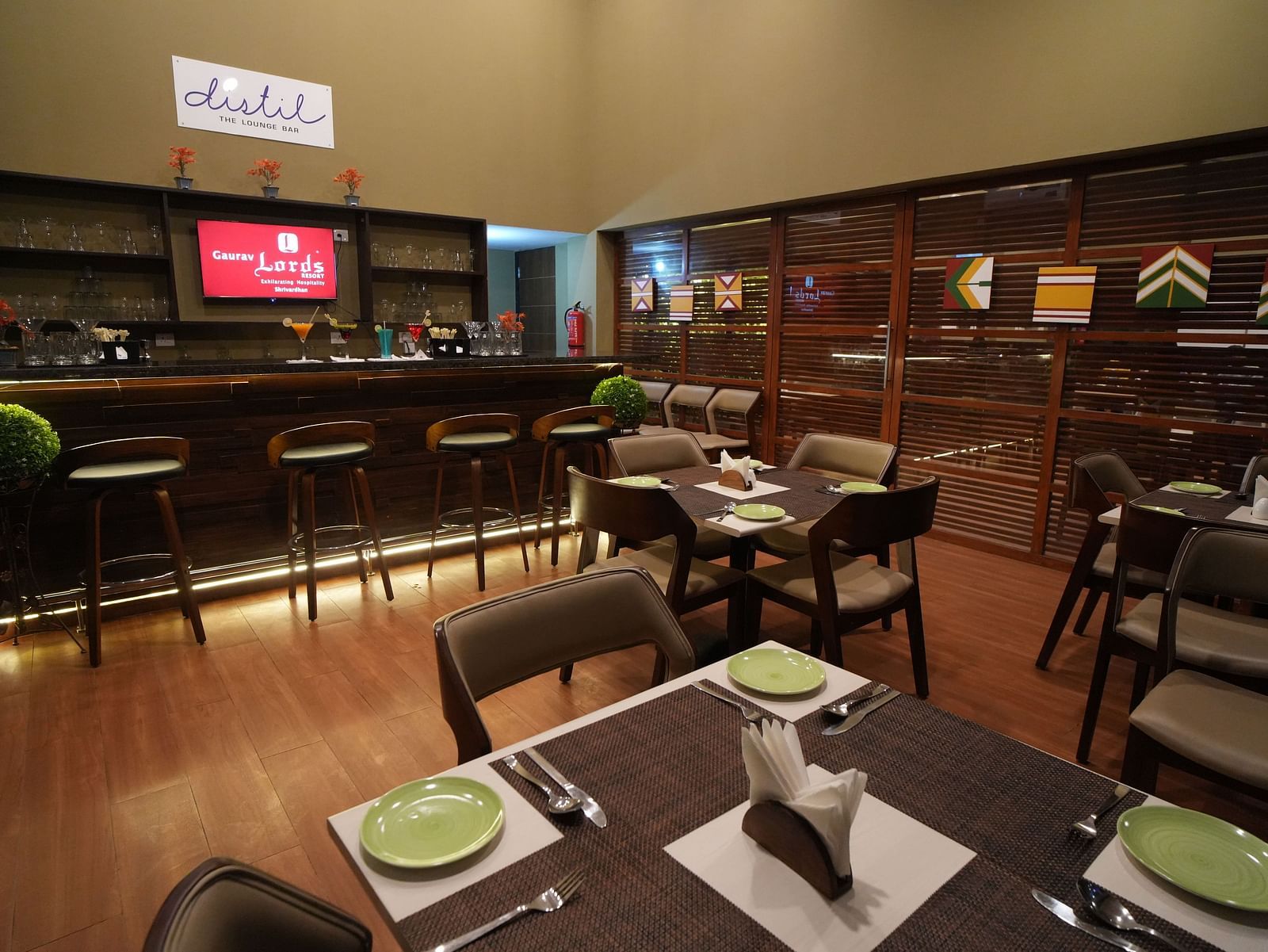 A warm, modern lounge bar with wooden flooring, neatly set dining tables, high bar stools, a well-stocked bar counter, and decor featuring colourful wall art - Gaurav Lords Resort Shrivardhan