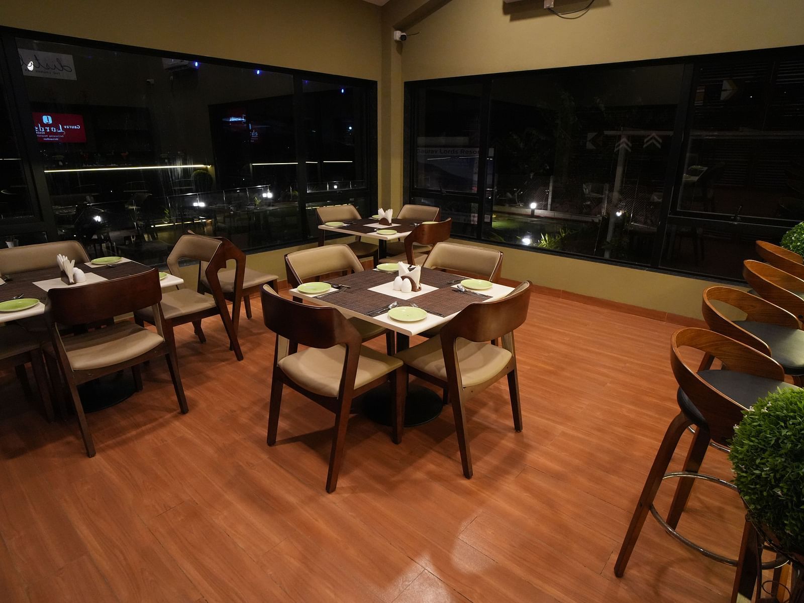 Indoor dining area with wooden flooring, cushioned chairs, neatly set tables, and floor-to-ceiling windows offering a night view, adjacent to a row of bar stools - Gaurav Lords Resort Shrivardhan