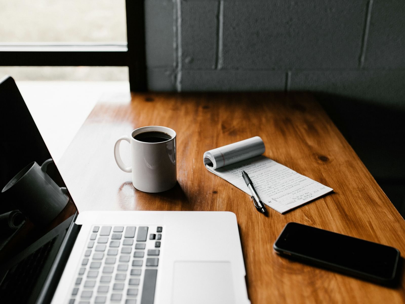 A laptop, smartphone, coffee cup, and notebook placed on a wooden desk
