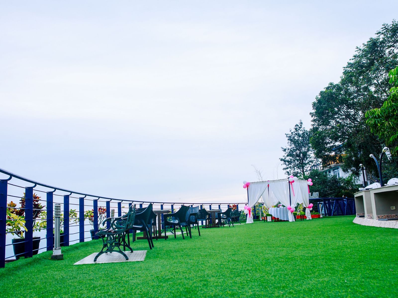 Open-air event space at GReaT Trails Yercaud by GRT Hotels, a corporate hotel in Yercaud, featuring a lush green lawn, outdoor seating, a decorated canopy with white drapes and pink balloons, and scenic surroundings