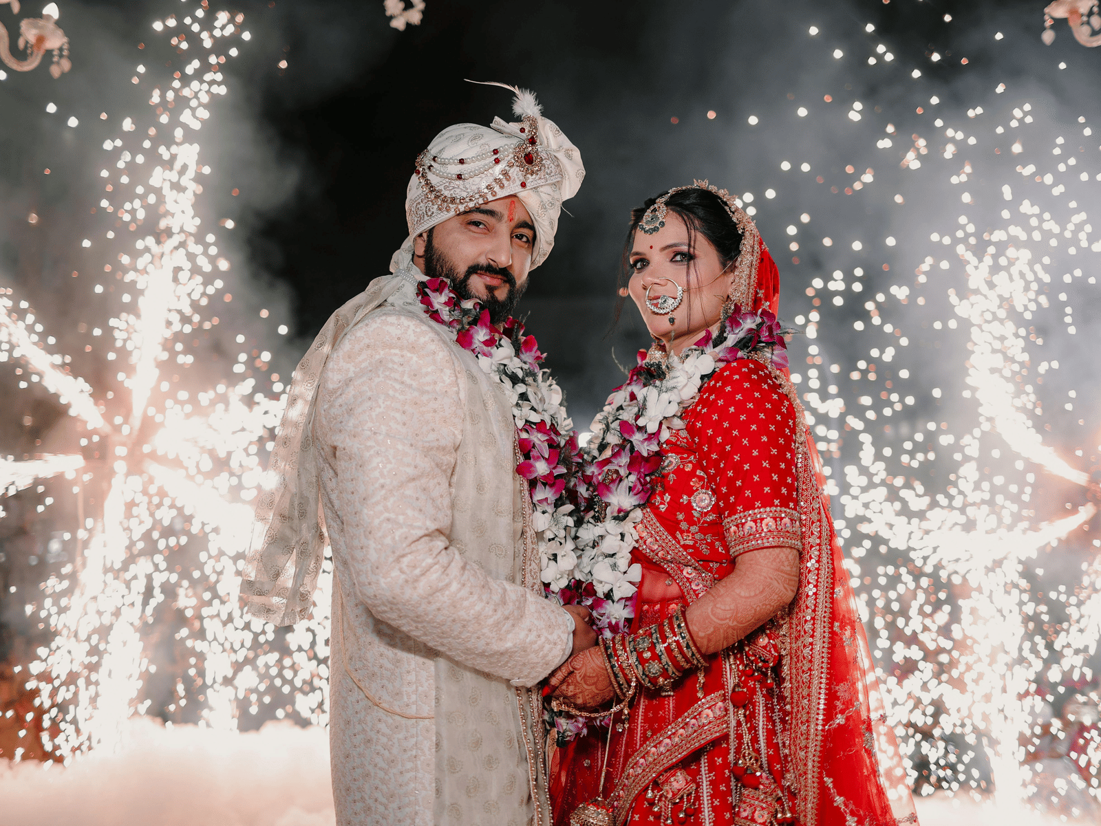 A bride and groom posing with fireworks in the background during a grand wedding photoshoot.