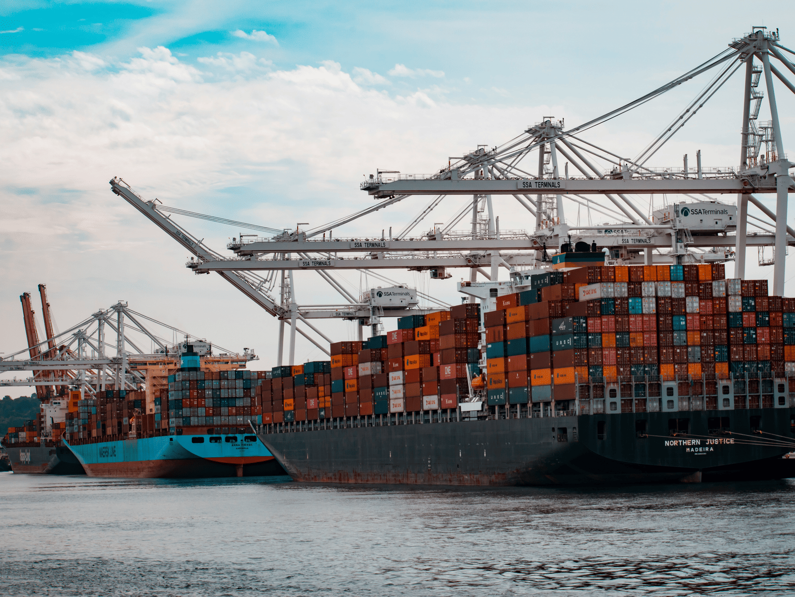 Several massive container ships laden with brightly coloured containers are docked beneath an array of towering port cranes against a cloudy blue sky.