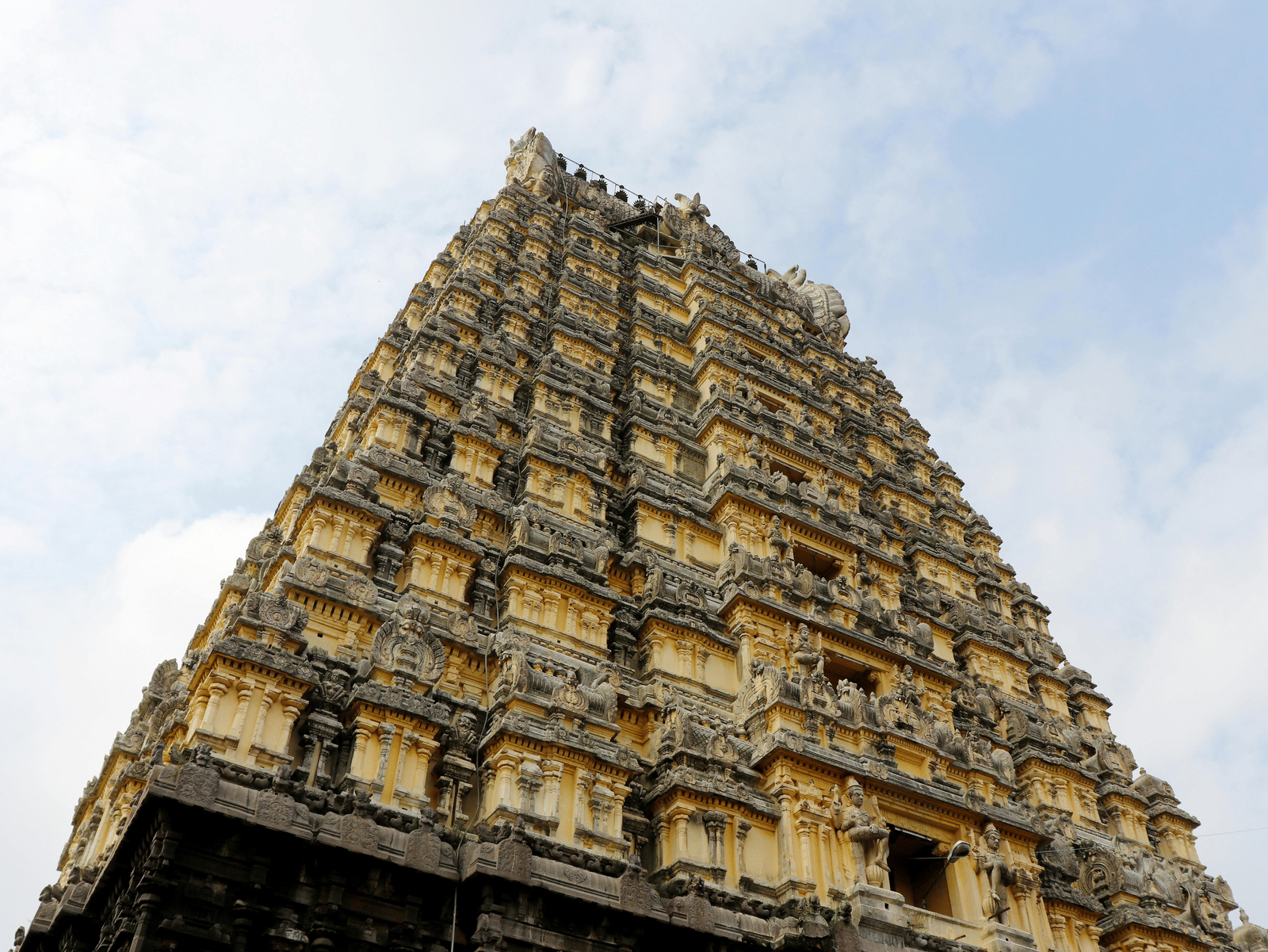 Close-up, low-angle view of a towering, pyramidal stone temple gateway (gopuram) against a cloudy sky.