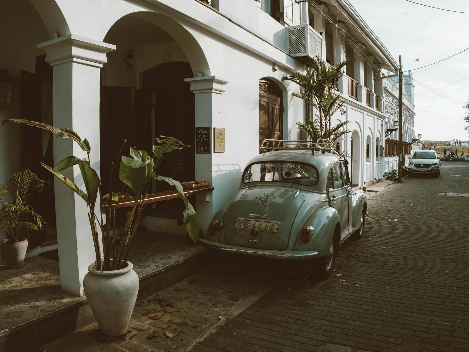 Colonial-style street lined with white arches, potted plants, and a parked vintage car, capturing the charm and heritage of an old town.