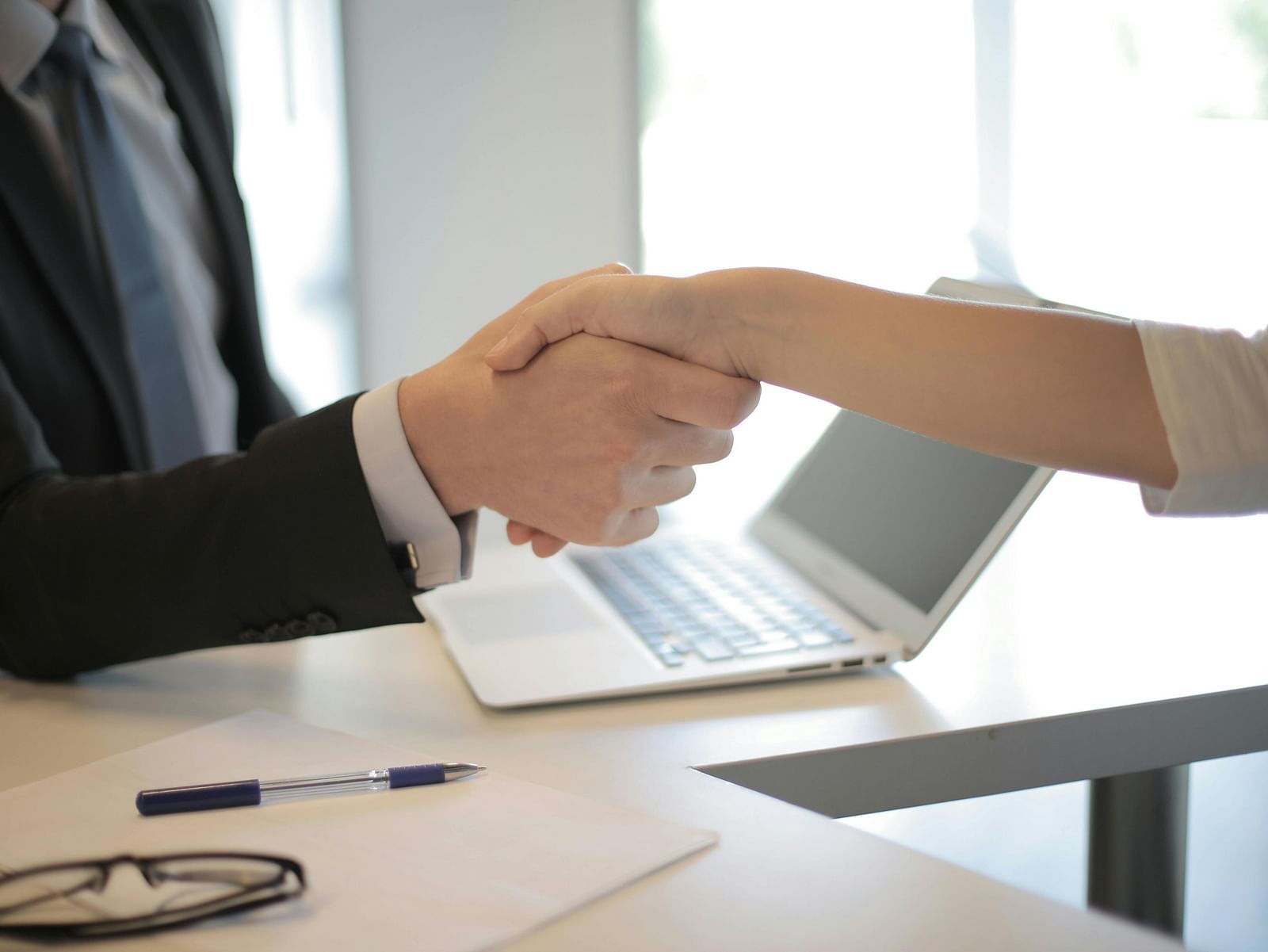 Two people in professional attire are shaking hands across a desk, with a laptop and documents visible, signifying a business agreement or greeting.