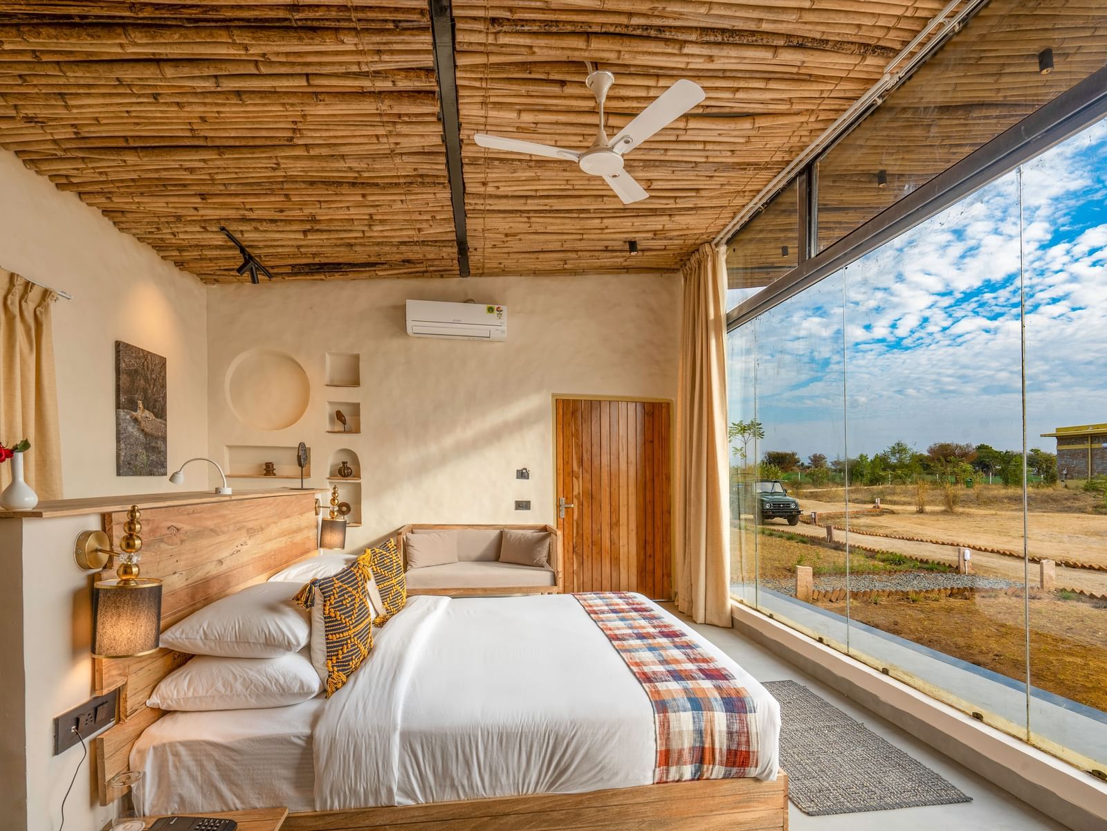 An image of a wooden furnished bedroom featuring a double size bed, a bedside table with lamp and socket, a sofa, some decorative elements in the room, and a large window from where outdoor scenery can be enjoyed - Kurja Jawai