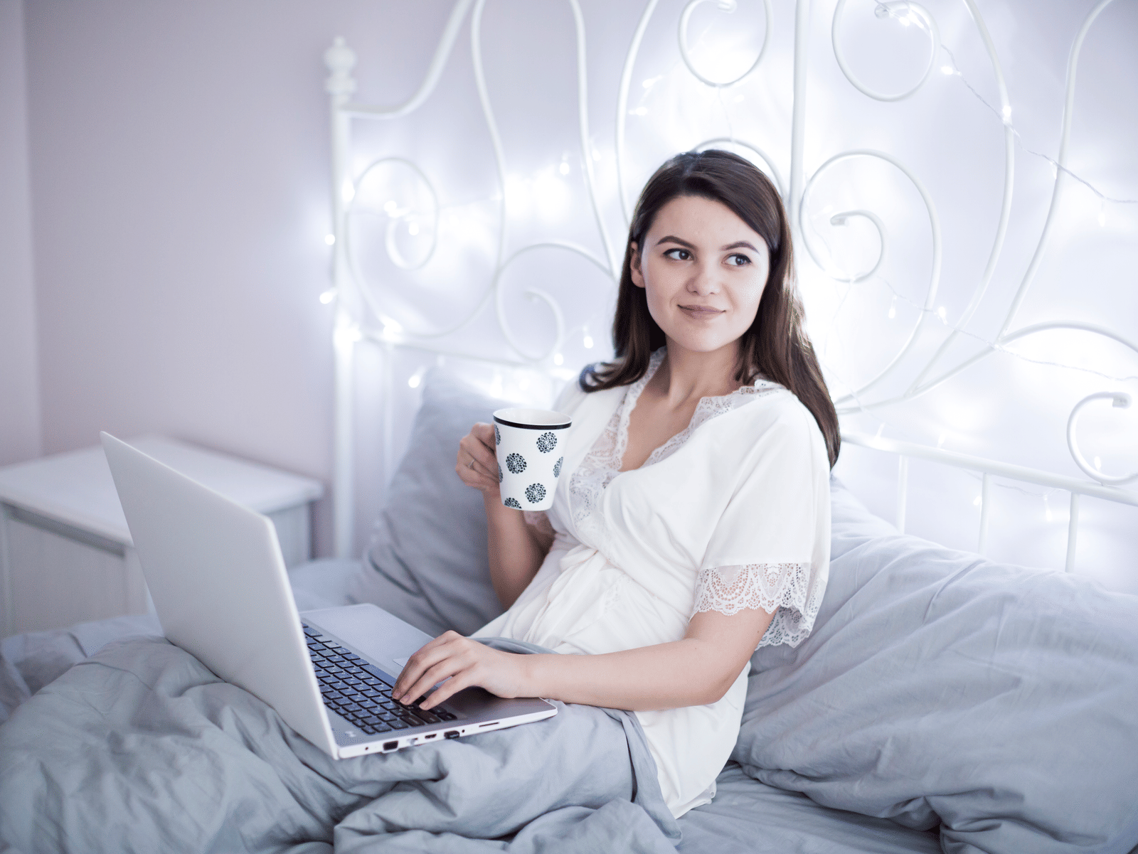 A smiling girl sitting with a cup in her hand and a laptop on her lap on a comfortable mattress with fancy LED lights on the metal head board.